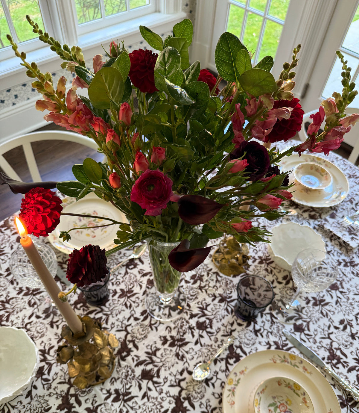 A large floral centerpiece with red roses, burgundy calla lilies, and green foliage arranged in a clear glass vase on a decorated dining table with vintage-style plates, a lit candle, and glassware.
